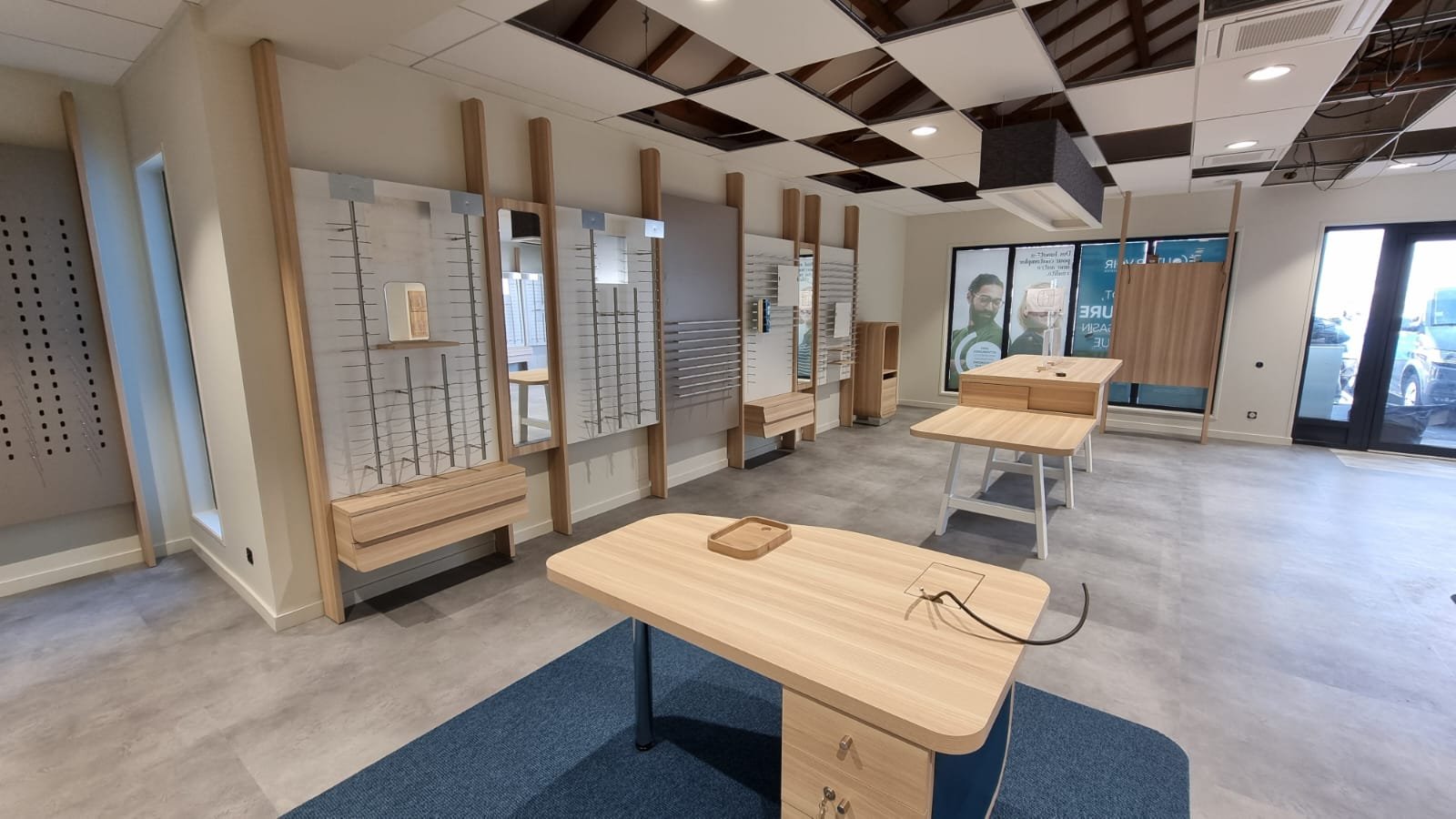 Après : magasin d'optique agencé avec murs de montures, miroirs, bureaux de conseil en bois et tapis bleu
